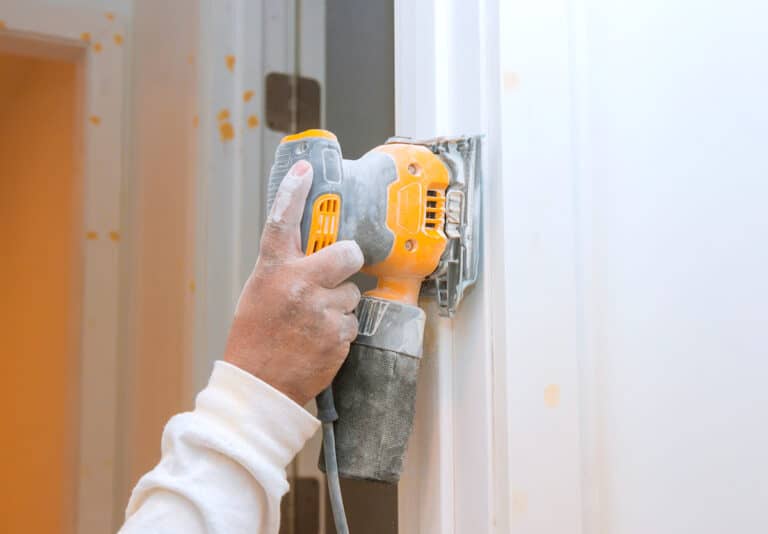 A dusty hand holding a yellow and grey sheet sander against the edge of a white painted door frame