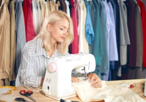 Best Sewing Machines For Clothes - The Creative Folk Sewing at a machine beside a rail of clothes
